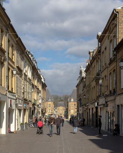 A pedestrianized urban street lined with multi-story stone buildings housing retail shops, featuring overhead tram or trolleybus wires and a distant classical building at the terminus.