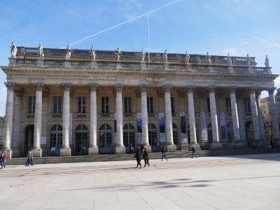 The image shows the grand neoclassical facade of the Grand Théâtre de Bordeaux, featuring a colonnade of Corinthian columns, statues atop the roofline, arched windows and entrances, and banners hanging between columns; people are walking on the pa