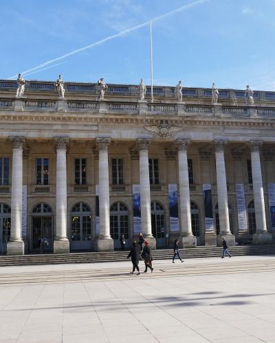 The image shows the grand neoclassical facade of the Grand Théâtre de Bordeaux, featuring a colonnade of Corinthian columns, statues atop the roofline, arched windows and entrances, and banners hanging between columns; people are walking on the pa