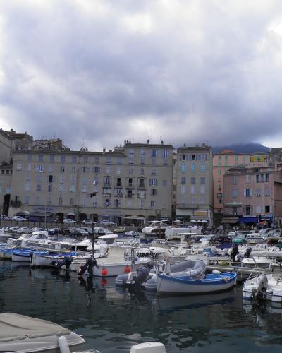A view of a crowded harbor filled with small pleasure boats and yachts, backed by a row of multi-story buildings in pastel colors (yellow, pink, beige) and a mountain peak visible in the distance under a cloudy sky.