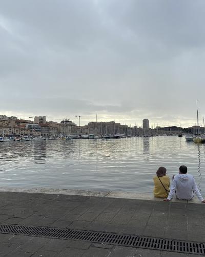 The image shows a wide, modern harbor with a paved promenade in the foreground, numerous docked sailboats and yachts, and a backdrop of multi-story urban buildings under an overcast sky.