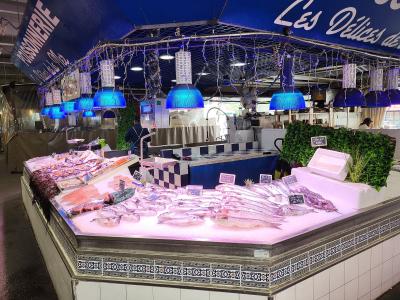 A fishmonger's stall inside a covered market, featuring fresh seafood on ice under blue heat lamps, with a sign reading "POISSONNERIE" and another partially visible sign saying "Les Délices de...".
