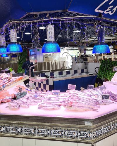 A fishmonger's stall inside a covered market, featuring fresh seafood on ice under blue heat lamps, with a sign reading "POISSONNERIE" and another partially visible sign saying "Les Délices de...".