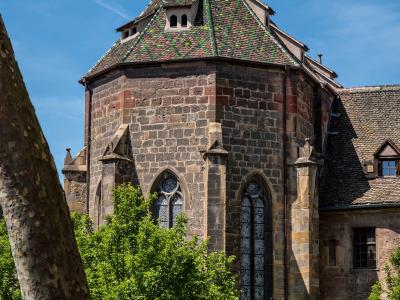 The image shows the exterior of a historic stone building with a distinctive octagonal tower featuring a steep, patterned tile roof and Gothic arched windows, set within a green park-like area with visitors on benches.