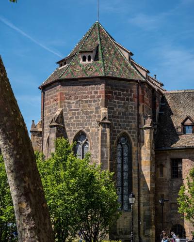 The image shows the exterior of a historic stone building with a distinctive octagonal tower featuring a steep, patterned tile roof and Gothic arched windows, set within a green park-like area with visitors on benches.