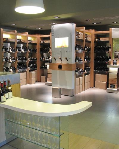 The image shows the modern interior of a wine shop or tasting room, featuring wooden shelving units stocked with wine bottles, a central dispensing station, a tasting counter with glasses, and signage including the word "SELECTION" and "Les Essent