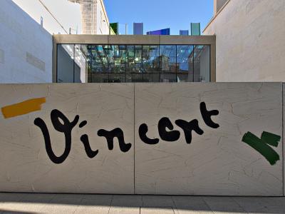 A low, textured white wall with the name "Vincent" painted in large black brushstrokes, flanked by yellow and green paint marks, with a modern glass building and colorful glass panels visible behind it.