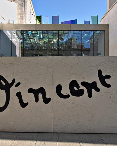 A low, textured white wall with the name "Vincent" painted in large black brushstrokes, flanked by yellow and green paint marks, with a modern glass building and colorful glass panels visible behind it.