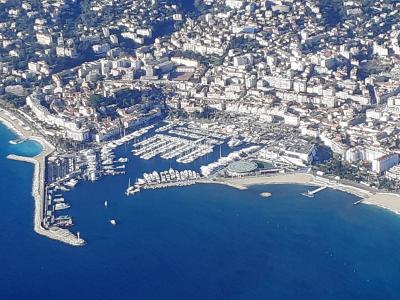 An aerial view of a large, densely packed marina filled with numerous white yachts and boats, situated along a curved coastline backed by a densely built-up urban area with white buildings and terracotta roofs, featuring a prominent breakwater and