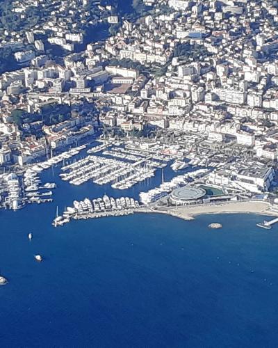 An aerial view of a large, densely packed marina filled with numerous white yachts and boats, situated along a curved coastline backed by a densely built-up urban area with white buildings and terracotta roofs, featuring a prominent breakwater and