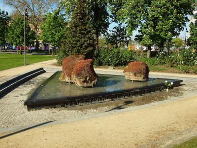 The image shows a modern park fountain featuring three large, moss-covered rock formations set within a rectangular black basin, with cobblestone pathways and green trees in the background.