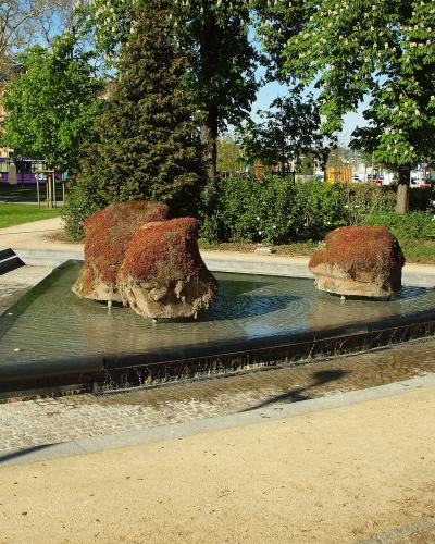 The image shows a modern park fountain featuring three large, moss-covered rock formations set within a rectangular black basin, with cobblestone pathways and green trees in the background.