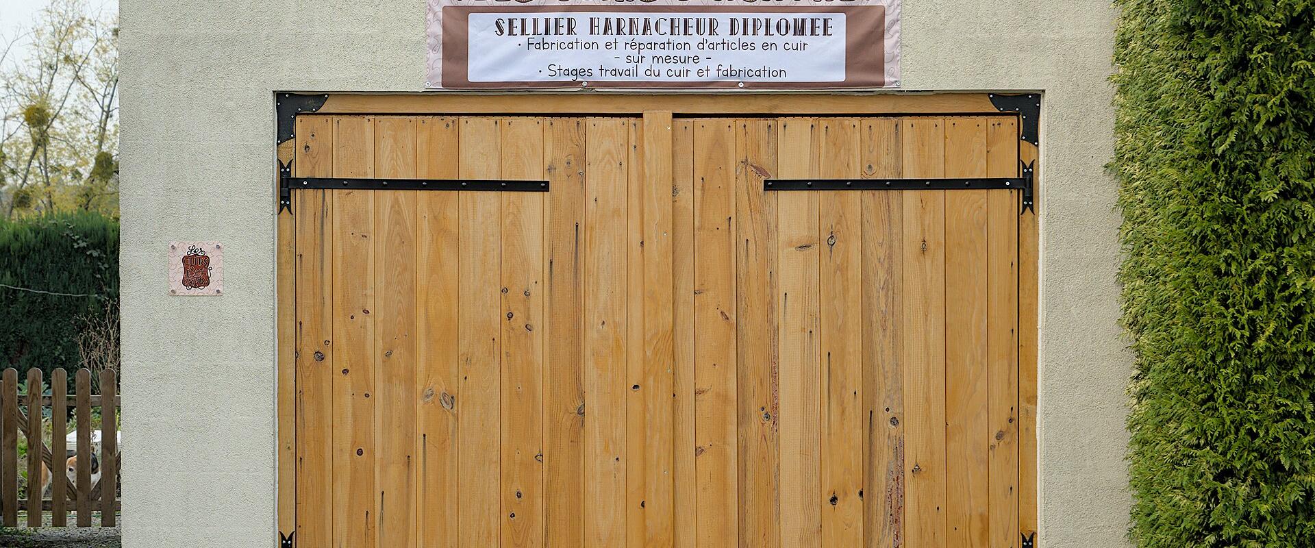 The image shows the exterior of a small workshop building with large wooden double doors, a sign reading "LES CUIRS D'AGATHE" and "SELLIER HARNACHEUR DIPLOMEE," indicating leatherwork and saddlery services, including craft workshops ("Stages trava