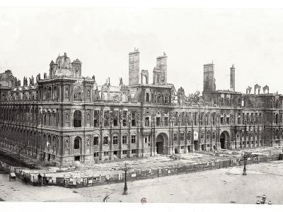 A black-and-white historical photograph showing a large, ornate, multi-story building with extensive fire damage, collapsed roofs, exposed chimneys, and debris-strewn grounds, consistent with the aftermath of the Paris Commune in 1871; no visible