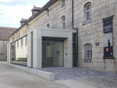 The image shows the modern glass-and-concrete entrance extension attached to a historic stone building, with a vertical banner on the right wall reading "MUSÉE DE LA RÉSISTANCE ET DE LA DÉPORTATION" and a small blue plaque below it indicating "Bes