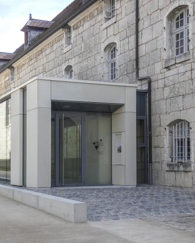 The image shows the modern glass-and-concrete entrance extension attached to a historic stone building, with a vertical banner on the right wall reading "MUSÉE DE LA RÉSISTANCE ET DE LA DÉPORTATION" and a small blue plaque below it indicating "Bes