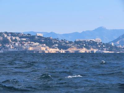 A seascape view from the water looking toward a hilly, densely built coastline with white buildings and cliffs under a clear blue sky, with distant mountains in the background; no visible signage or specific landmarks identifying the Corniche Kenn