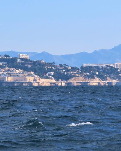 A seascape view from the water looking toward a hilly, densely built coastline with white buildings and cliffs under a clear blue sky, with distant mountains in the background; no visible signage or specific landmarks identifying the Corniche Kenn