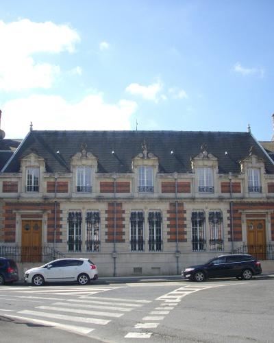 A large, ornate building with red brick and stone detailing, mansard roofs, and dormer windows, situated on a street corner with parked cars and a crosswalk.