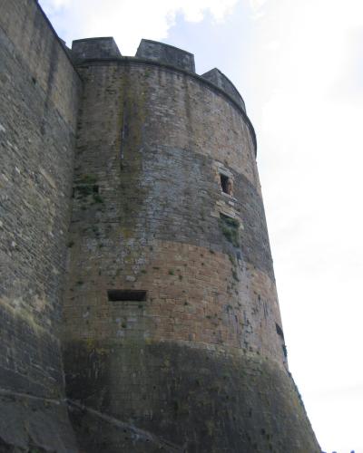 A low-angle shot of a large, cylindrical stone fortification tower with crenellations at the top, featuring small windows and weathered masonry with vegetation growing on its surface.