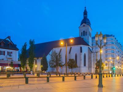 The image shows the Église Saint-Maurice in Annecy, France, captured at dusk with its white stone facade, Gothic arched windows, and a prominent bell tower topped with a dark spire, situated in a paved public square with streetlights and modern bu