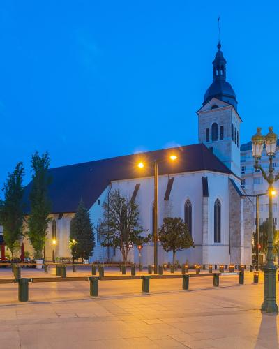 The image shows the Église Saint-Maurice in Annecy, France, captured at dusk with its white stone facade, Gothic arched windows, and a prominent bell tower topped with a dark spire, situated in a paved public square with streetlights and modern bu