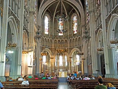 The image shows the interior nave of a large Gothic-style basilica with high vaulted ceilings, stained glass windows, ornate chandeliers, and rows of wooden pews filled with visitors facing an altar at the far end.