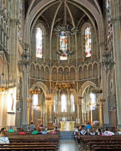 The image shows the interior nave of a large Gothic-style basilica with high vaulted ceilings, stained glass windows, ornate chandeliers, and rows of wooden pews filled with visitors facing an altar at the far end.