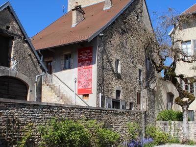 A stone building with a red banner reading "1855/2005" and text referencing Gustave Courbet and Realism, situated behind a low stone wall with greenery and a grassy foreground under a clear blue sky.