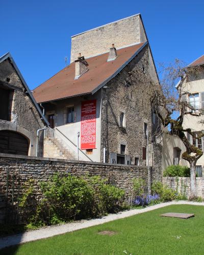 A stone building with a red banner reading "1855/2005" and text referencing Gustave Courbet and Realism, situated behind a low stone wall with greenery and a grassy foreground under a clear blue sky.