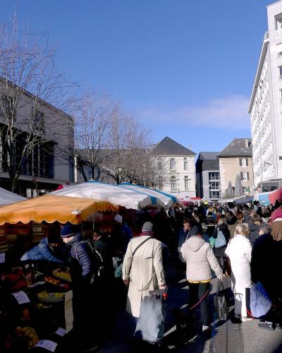 A crowded outdoor street market with colorful vendor stalls and awnings, set between modern and traditional buildings under a clear blue sky.