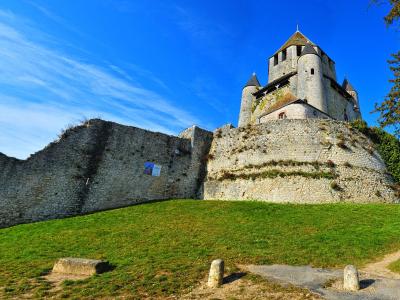 A low-angle view of a medieval stone fortress with conical-roofed towers and high curtain walls situated atop a grassy mound under a blue sky, with a small poster visible on the lower wall.