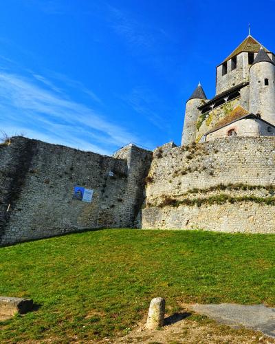 A low-angle view of a medieval stone fortress with conical-roofed towers and high curtain walls situated atop a grassy mound under a blue sky, with a small poster visible on the lower wall.