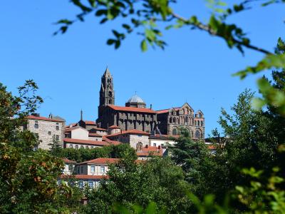 A large, dark-stoned Romanesque cathedral with a prominent bell tower and red-tiled roofs, viewed through green foliage against a clear blue sky, situated on a hill above surrounding buildings.