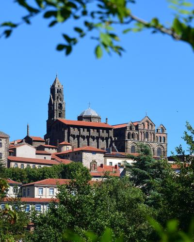 A large, dark-stoned Romanesque cathedral with a prominent bell tower and red-tiled roofs, viewed through green foliage against a clear blue sky, situated on a hill above surrounding buildings.