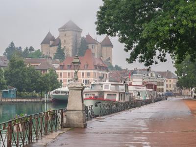 A misty, overcast view of a stone castle with conical-roofed towers perched on a hill overlooking a canal lined with boats and a wet promenade in the foreground.