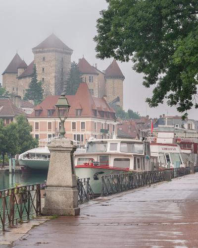 A misty, overcast view of a stone castle with conical-roofed towers perched on a hill overlooking a canal lined with boats and a wet promenade in the foreground.
