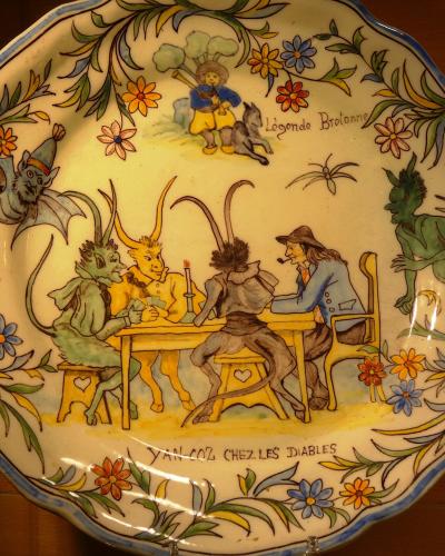 A close-up photograph of a decorative ceramic plate featuring a painted scene of a man playing cards with three demons around a table, surrounded by a floral border and inscribed with "Légende Bretonne" and "YAN COZ CHEZ LES DIABLES".