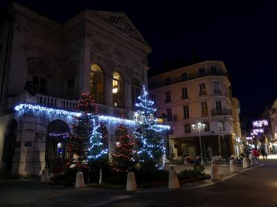 A night view of a classical stone building with arched windows and a pediment, decorated with Christmas trees and blue/white lights, with a sign reading "MISE EN SCENE" on an adjacent building.