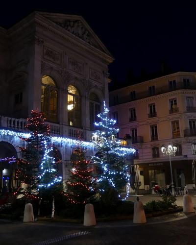 A night view of a classical stone building with arched windows and a pediment, decorated with Christmas trees and blue/white lights, with a sign reading "MISE EN SCENE" on an adjacent building.