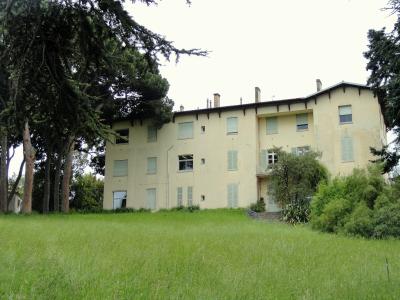 A large, pale yellow villa with shuttered windows and a tiled roof, set on a grassy slope with mature trees to the left and shrubbery to the right; no visible signage or text.