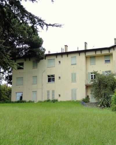 A large, pale yellow villa with shuttered windows and a tiled roof, set on a grassy slope with mature trees to the left and shrubbery to the right; no visible signage or text.