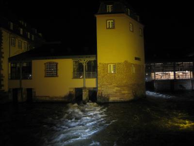 A night photograph of a yellow-walled building with a stone base situated on a riverbank, featuring a prominent cylindrical tower section and a sign reading "HOTEL" on its side, with water flowing rapidly in the foreground.