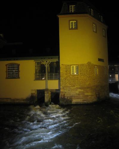 A night photograph of a yellow-walled building with a stone base situated on a riverbank, featuring a prominent cylindrical tower section and a sign reading "HOTEL" on its side, with water flowing rapidly in the foreground.