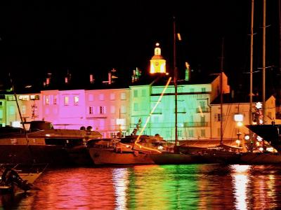 A night view of a harbor with colorful buildings and boats, featuring a prominent illuminated church tower in the background.