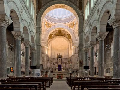 The image shows the interior nave and apse of a large church with Roman-Byzantine architecture, featuring massive granite columns, rounded arches, a central dome with mosaics, and an altar area with purple liturgical cloth; signage in the foregrou
