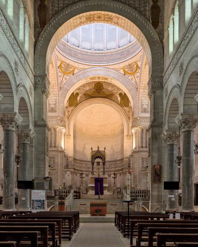 The image shows the interior nave and apse of a large church with Roman-Byzantine architecture, featuring massive granite columns, rounded arches, a central dome with mosaics, and an altar area with purple liturgical cloth; signage in the foregrou