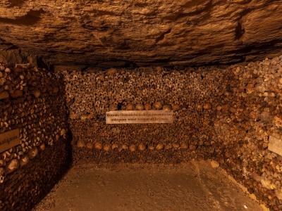 1. A large, cylindrical ossuary structure constructed from neatly stacked human femurs and skulls, situated within a rough-hewn limestone tunnel with a stone cross visible on the left wall.