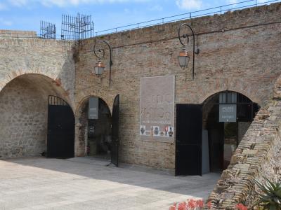 The image shows the exterior entrance of a historic brick and stone fortress with arched gateways, featuring signage that reads "MUSÉE D'ARCHÉOLOGIE ANTIBES" and a large poster displaying archaeological artifacts.