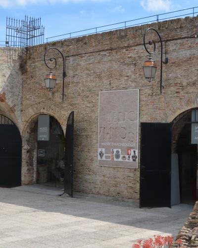 The image shows the exterior entrance of a historic brick and stone fortress with arched gateways, featuring signage that reads "MUSÉE D'ARCHÉOLOGIE ANTIBES" and a large poster displaying archaeological artifacts.